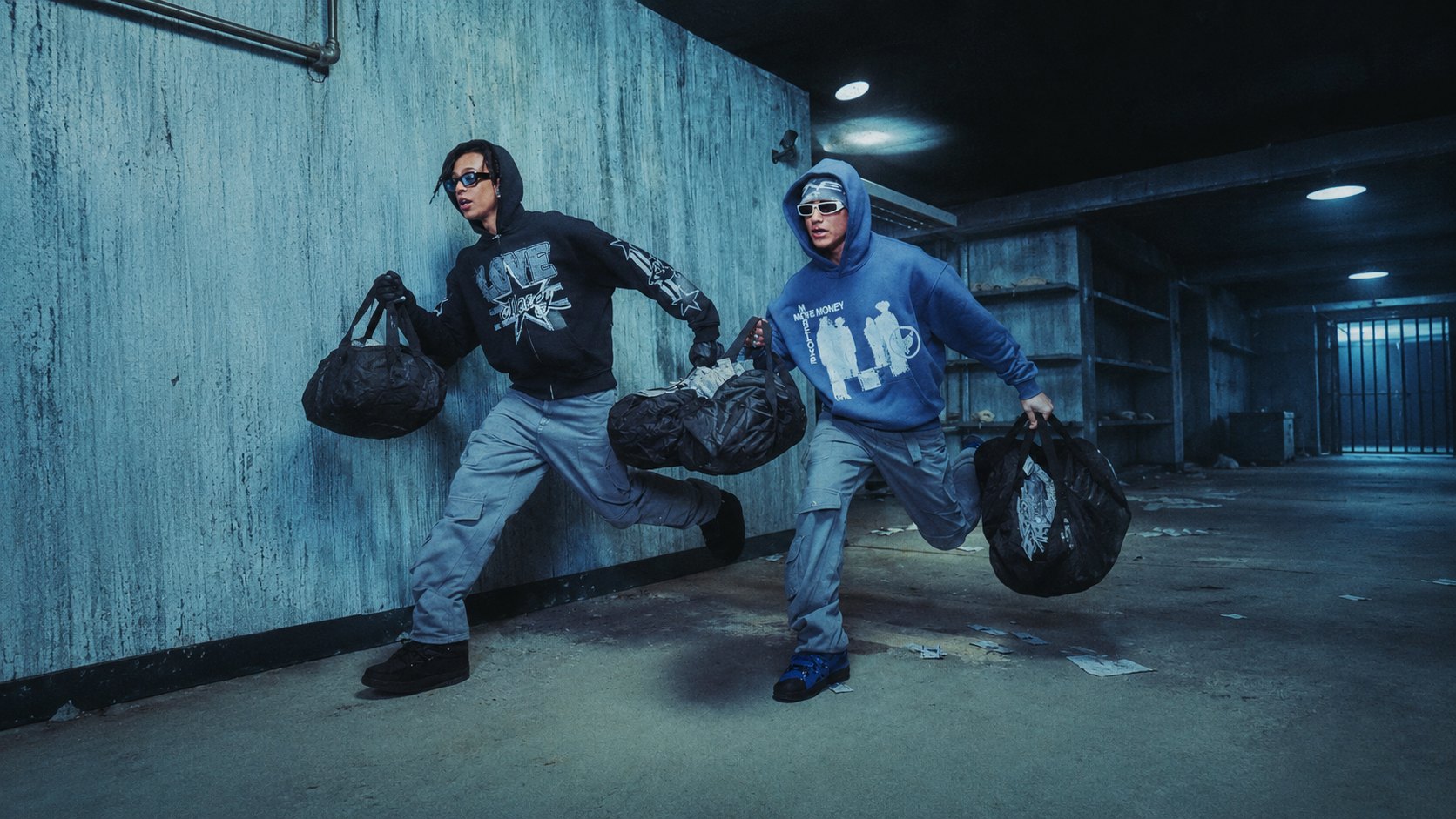 Two young men in hoodies and cargo pants run through a concrete corridor, a black duffel bag in each hand